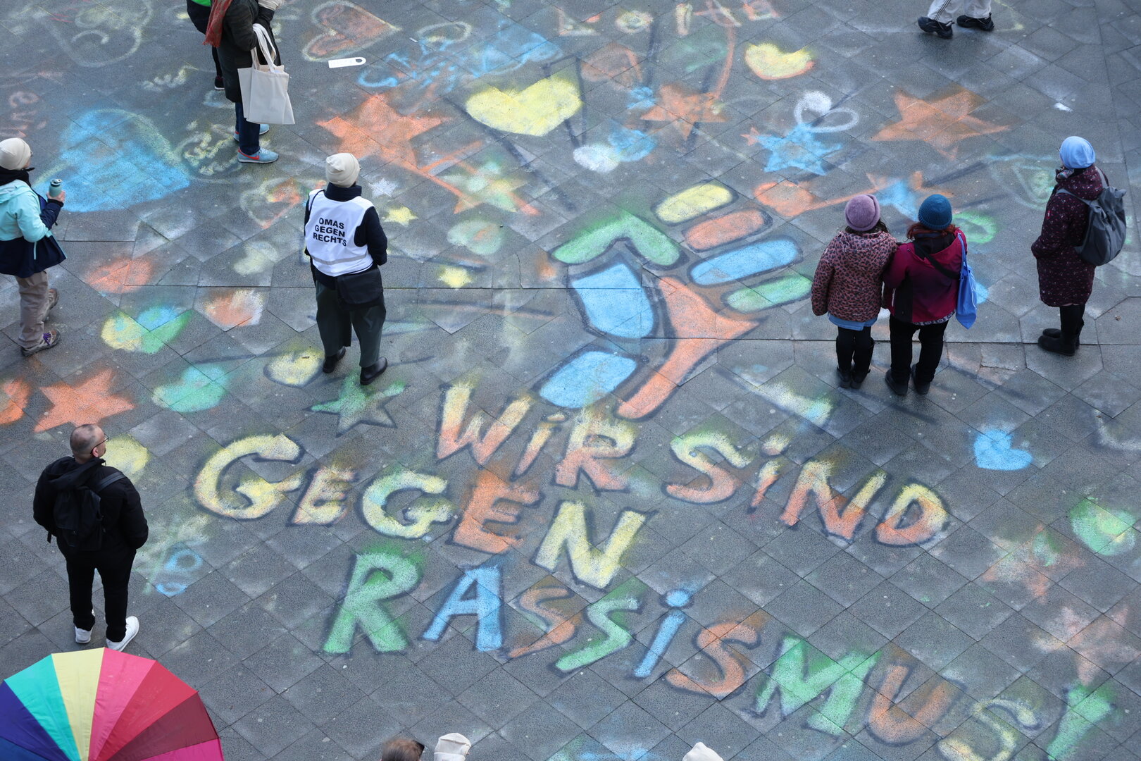 Menschen auf einem Platz mit Kreidekunst: 'WIR SIND GEGEN RASSISMUS' und ein Protest-Faustsymbol.