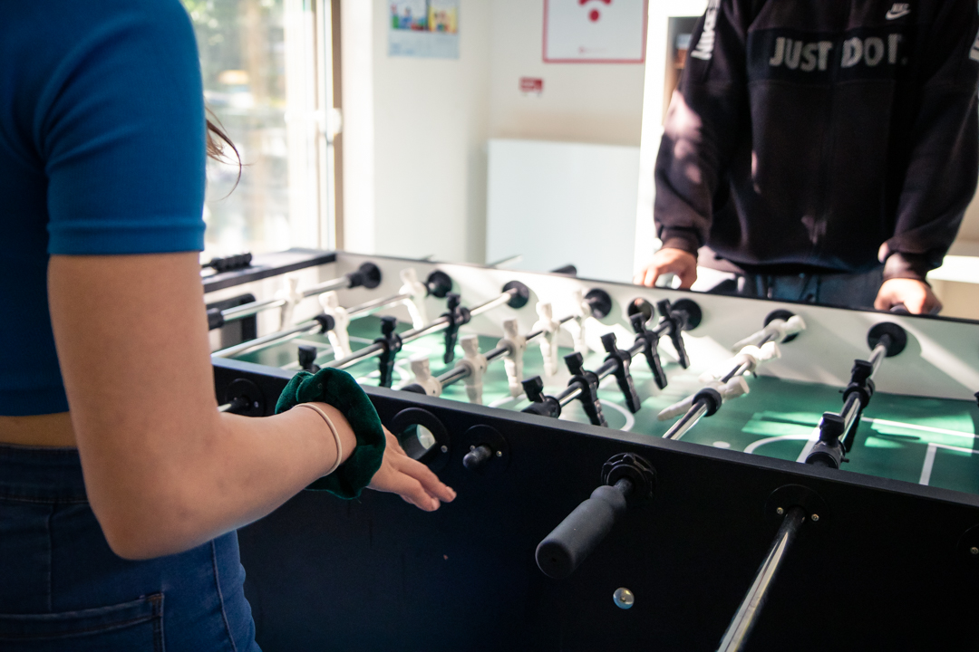 Zwei Personen spielen Kicker. Eine Person in blauem Oberteil mit Haargummi, die andere in schwarzem Hoodie.