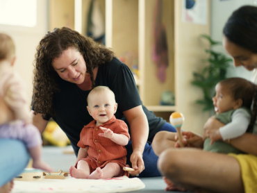 In einer Krabbelgruppe sitzt ein Baby auf einer Spielmatte, während eine pädagogische Fachkraft und weitere Erwachsene die Kinder beim gemeinsamen Spielen begleiten.